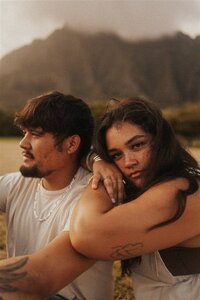 A couple sits close together on the ground with a mountainous backdrop in Oahu, Hawaii, capturing a quiet and intimate moment at sunset.