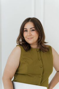 Confident woman with medium brown hair in an olive green top smiling while holding a laptop.
