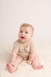 6 month old baby girl sitting up and smiling at the camera. She is wearing a crocheted beige romper and sitting on a beige carpet at a portland baby photography studio.