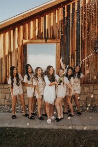 A wedding party celebrates at a ranch wedding in Dallas, Texas. The group, dressed in coordinated outfits, gathers outside a rustic wooden barn as they pop a bottle of champagne, capturing the fun and excitement of the day.