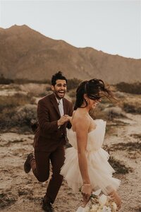 A couple joyfully runs through a desert landscape in Palm Springs, California, during their elopement. One partner wears a ruffled, strapless dress, while the other is dressed in a suit, both laughing as the wind blows through their hair.