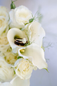 A close up of a diamond ring and a black wedding band nestled inside white flowers