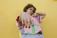 A young person with curly hair, poses with their right arm outstretched toward the camera, black nail polish visible on their fingertips. They look down their arm, toward the viewer, with a confident expression. Their other arm is bent, so their hand rests under their chin. The wall behind them is yellow, and they are wearing a brightly colored button-down shirt with jeans.