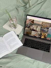 Open book, open laptop with images displayed and glass of water on top of green duvet.