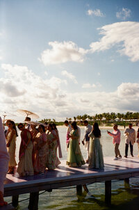 Guests at an Indian wedding in Playa del Carmen, Mexico, walking across a picturesque walkway over the water toward the ceremony site, dressed in vibrant Indian wedding attire and carrying parasols under the tropical sun.