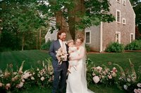 Intimate backyard wedding on Cape Cod with bride and groom posing in front of meadow floral arrangement holding baby wearing flower crown.