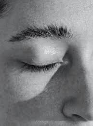 A black and white close-up portrait of the corner of an eye, showcasing eyelashes, eyebrows, and a slight smile. The face is illuminated by soft natural light from above, emphasizing the serene expression and adding depth to the portrait. The minimalist composition highlights the calmness and tranquility in the subject’s gaze, reflecting the elegance of subtle beauty. This image, ideal for high-end branding, can be used in creative branding design, luxury brand identity, or custom website design to evoke sophistication and emotional connection.