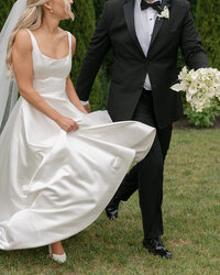 Bride in a white gown and groom in a black tuxedo joyfully walking on grass. She holds her dress, and he carries a bouquet of white flowers.