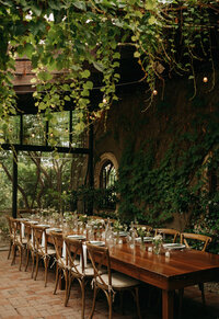 Romantic outdoor wedding reception table under vines and string lights, set for an intimate elopement dinner with wooden chairs and lush greenery.