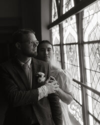 Bride and groom standing by a stained glass window, holding each other in soft light, captured by Rainstorm Photo & Video in a timeless, documentary wedding style.