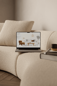 Laptop on a beige bouclé sofa, displaying a sample board for a living room design, with books and a cup of coffee placed in the foreground on the sofa.