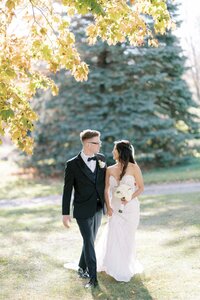 Newlywed couple walking hand in hand in a sunlit garden, bride in white dress and groom in classic black suit