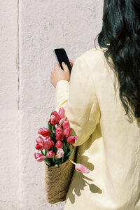 A woman wearing white on her phone holding a bag of pink tulips 