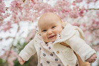 A baby is held up with cherry blossom behind her. She smiles at the camera during the blossom photo shoot in Bushy Park.