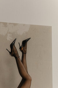  A close-up side view of a person's lower legs in black tights and heels, lifted in the air with a slight bend at the knees. The legs are positioned against a white wall with a beige canvas in the background.