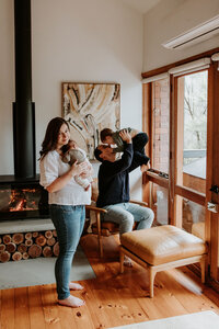Mum holds newborn baby while dad plays with toddler in background of lunge room at in home photography session