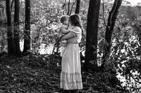 Black and white photo of a mom holding a toddler while they touch foreheads in a forested area.