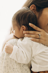 A mother affectionately holding her baby close, with the infant's head resting on her shoulder. the mother's hand gently supports the baby's head. soft, warm lighting enhances the tender moment.