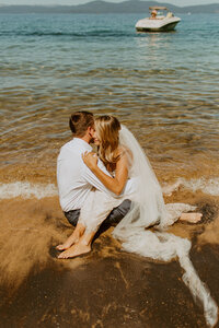 Beachside elopement, couple embraced, bride sitting in lap, couple in water on beach shore
