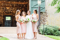 A bride stands in her dress in a patio laughing with her bridesmaids holding all their bouquets