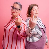 Two people, Pippa & Lucy, stand back-to-back in a studio, smirking as they hold up their middle fingers toward the camera. Pippa on the left wears glasses, a red-and-pink striped blouse, and jeans; Lucy on the right wears a black-and-white striped top with dusty-purple dungarees.