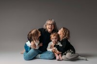 A group shot shows a grandmother surrounded by her three granddaughters, ages 2, 4, and 6. They are all wearing cozy sweaters, with the grandmother seated on the floor and holding the youngest granddaughter in her lap. The older granddaughters are gathered around, all smiling and enjoying the moment together in Mobile, AL Motherhood Photographer’s studio