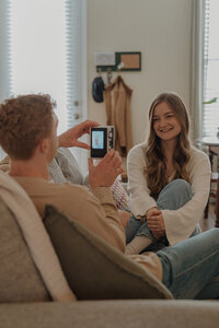 A man takes a photo of his girlfriend on a digital camera inside their home. She is smiling at him and he has his back to the camera.