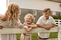three children stand on a fencepost at a farm, as they all laugh together and smile at the camera. The children wear neutral fall clothing and are laughing