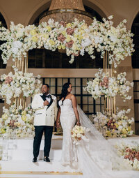 Bride and groom pose for portrait underneath lush white and gold floral installation