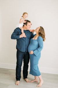 Toddler girl sits on dad's shoulders and smiles at mom during studio family maternity session
