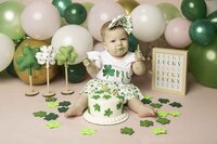 Little one joyfully smashing a birthday cake surrounded by scattered four-leaf clovers. The cake is decorated with vibrant frosting, and the baby’s face is filled with pure delight, as the lucky clovers add a touch of whimsy to the fun and playful moment