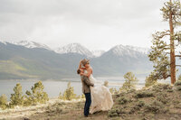 groom holding bride off the ground while they stand next to a lake with mountains in the background