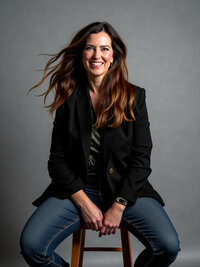 Missy Mayo is in a studio with a gray background and is sitting on a stool. Her hair is softly blowing and she is smiling at the camera.