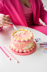 Kat holding a fork in front of a small cake with pink and yellow icing
