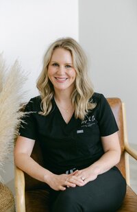 A woman with long brown hair wearing a black outfit sits smiling on a brown chair with her hands clasped, next to a decorative arrangement of light-colored pampas grass at a Maine medical aesthetics clinic.