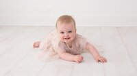 A baby is lying on the floor at Dandelion Studio in Moira, Northern Ireland, wearing a soft pastel dress. The baby has bright eyes and a curious smile, looking up at the camera in a sweet and natural moment.