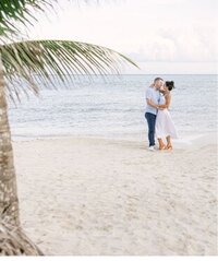 Palm tree overhanging a tropical beach shoreline where a couple embrace before their destination ceremony