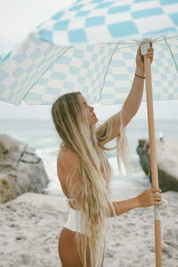 Blonde woman holding a blue-and-white checkered beach umbrella, standing on sandy shore with ocean rocks in the background — soft, summer coastal aesthetic.