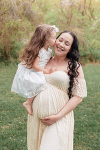 A pregnant mom holding her belly and getting a kiss from her daughter.