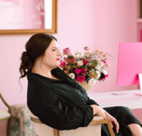 Confident woman with dark hair in a loose updo, wearing a black dress and sitting in a modern chair beside a pink desk setup, with a vibrant floral arrangement and soft pink walls in the background.