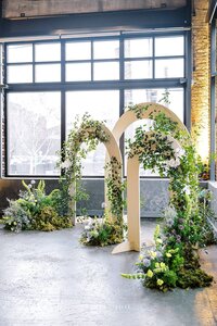 A posed wedding photo of a bride and groom standing and softly embracing each other. They are standing on a wooden deck with a black railing behind them. In focus, they have colorful floral arrangements on two pedestals, one floral arrangement on the ground, and a wedding bouquet in the bride's hand. The flowers are orange, blue, and yellow. The bride is wearing a champagne colored sequin dress that is floor length. The groom is wearing a black suit. In the background, there are large evergreen trees.