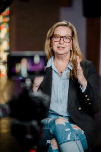 A woman in a blue collared shirt and jacket sits in front of a camera for an interview.