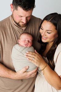 A couple stands together, smiling warmly, as they hold their newborn baby wrapped in a light blanket. The mother, wearing a patterned dress, gazes lovingly at the child, while the father, wearing a brown sweater, looks at both with pride. The background features soft, earthy tones that enhance the intimate, joyful atmosphere.
