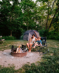 Near a campfire at Five Rivers Metroparks, a woman and child sit in chairs, sharing a cozy moment together.