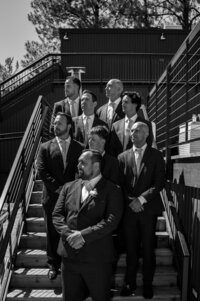 Candid black and white editorial-style photo of groomsmen in formal suits standing on outdoor stairs at River Run Lodge, captured during a stylish wedding celebration.