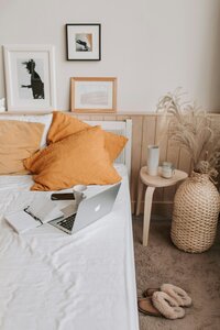 Bedroom showcasing framed art on the wall, arranged with orange pillows, Macbook Pro laptop, journal, iPhone, and mug on the bed with other decorative elements on the side of the bed