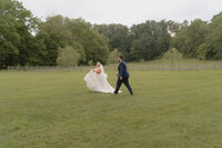 A groom chases his bride playfully in a field while she runs away and looks back at him at Hidden Meadows in Northeast, TN