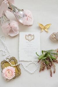 Gorgeous detail photo displaying the wedding invitation, rings, bride's shoes, embellished with pearls and florals.