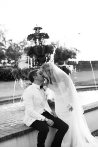 Editorial black and white wedding photo of bride and groom sitting at a fountain