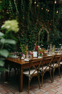 Romantic outdoor wedding reception table under vines and string lights, set for an intimate elopement dinner with wooden chairs and lush greenery at the Corson Building in Seattle, WA.